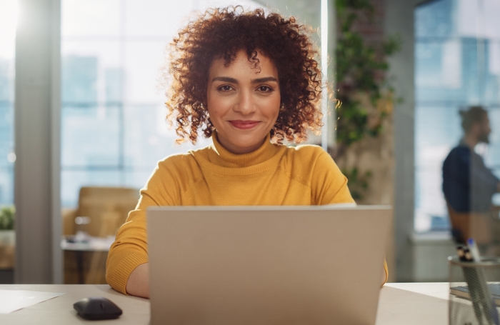 A woman sitting at a desk working at a laptop smiling to the camera LARGEjpg