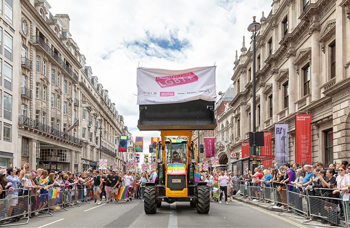 FeixandmerlinPrideImage 3jpg A rainbow painted digger with an Architecture LGBT+ banner and pride parade marchers behind it, with people waving rainbow flags on both sides of the road