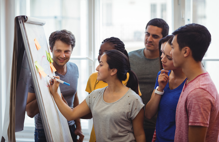 A group of people standing around a whiteboard with one taking the lead in conversation holding a pe