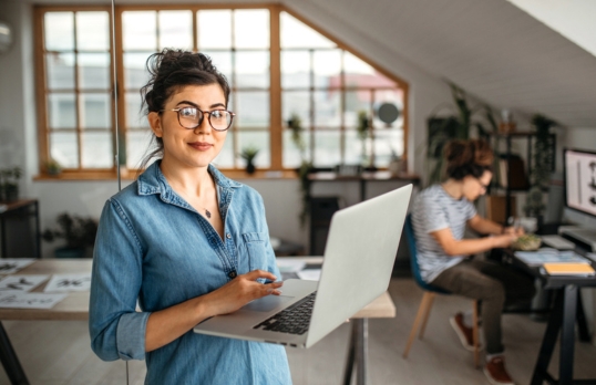 A young woman wearing glasses in an office holding a laptop GC SMALLjpg