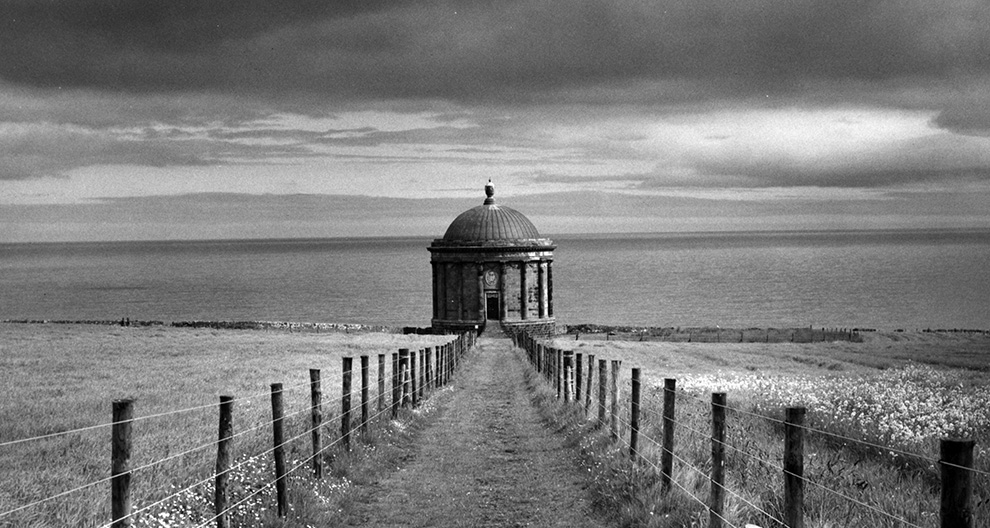 Black and white photograph of a dirt road with wire fence leading to a neoclassical temple building with columns and rounded roof, body of water in the background